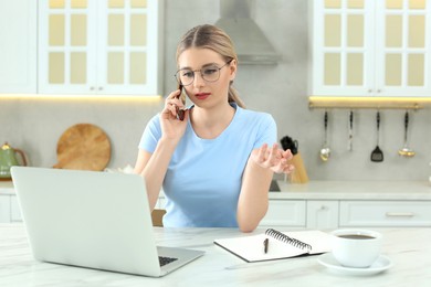 Home workplace. Woman talking on smartphone near laptop at marble desk in kitchen