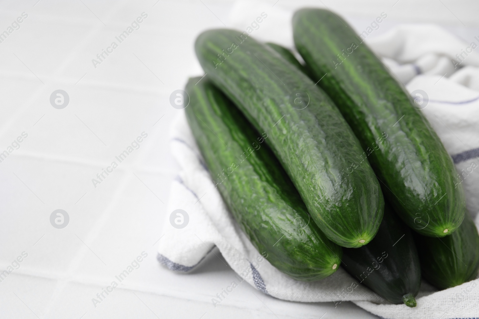 Photo of Fresh cucumbers and cloth on white tiled table, closeup. Space for text