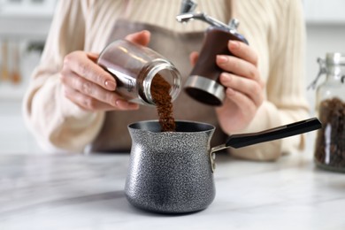 Photo of Woman pouring coffee powder from grinder into jezve at table indoors, closeup
