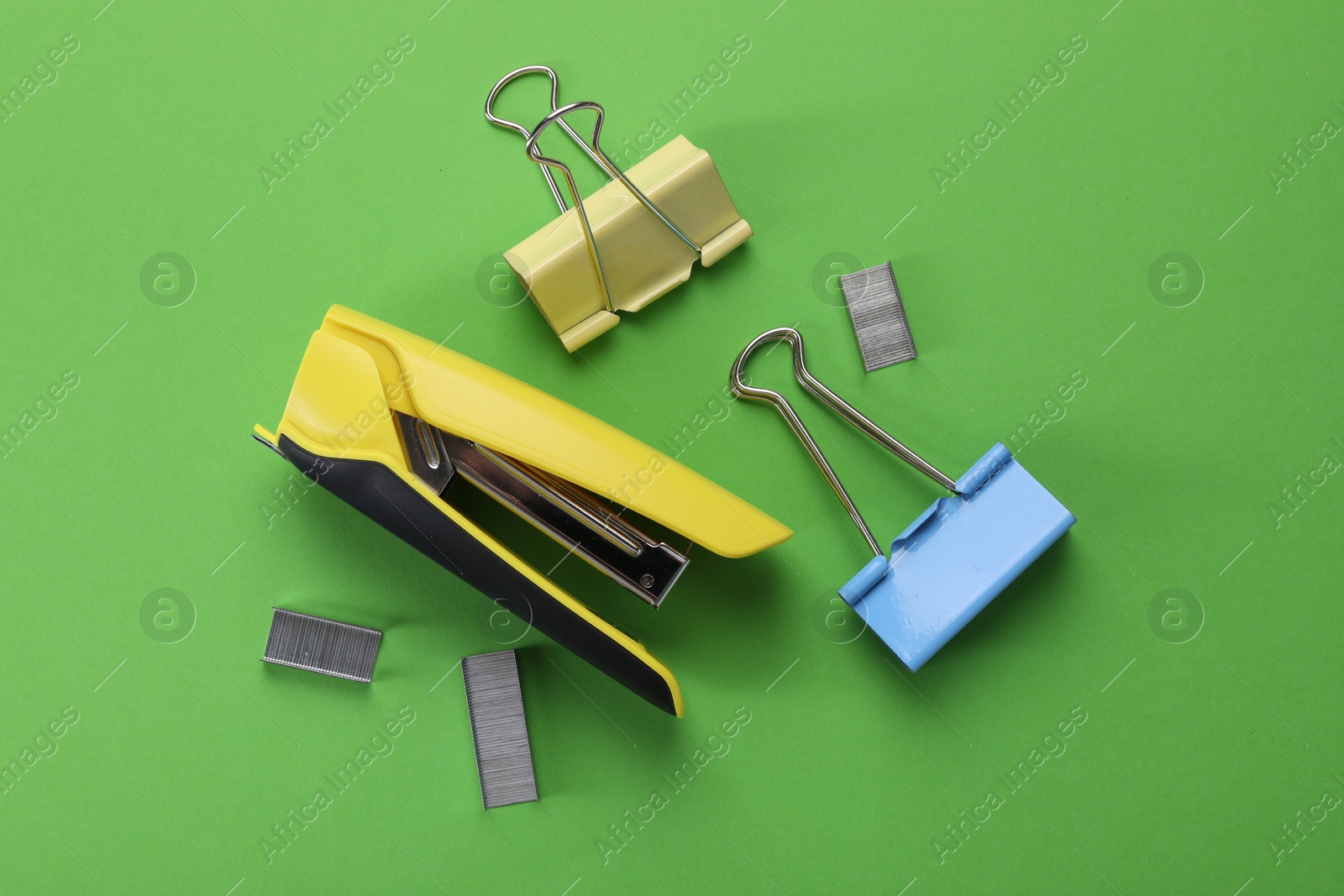 Photo of Yellow stapler with staples and binder clips on green background, flat lay