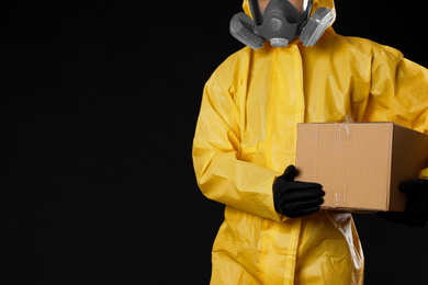 Photo of Man with cardboard box wearing chemical protective suit and space for text on black background, closeup. Prevention of virus spread