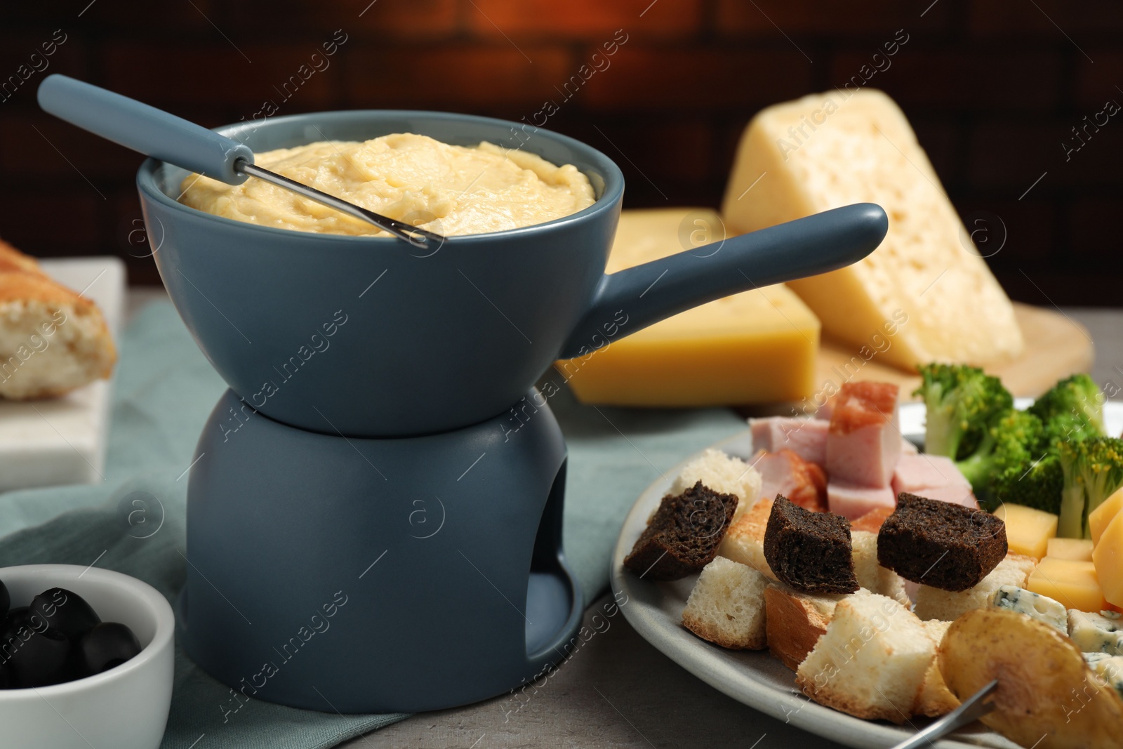 Photo of Tasty melted cheese in fondue pot, fork and snacks on grey table, closeup