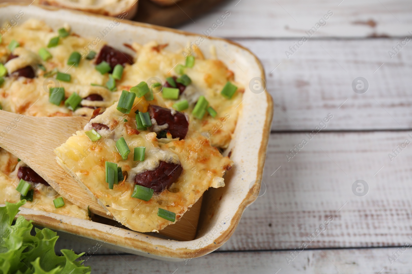 Photo of Taking piece of tasty sausage casserole from baking dish at white wooden table, closeup. Space for text