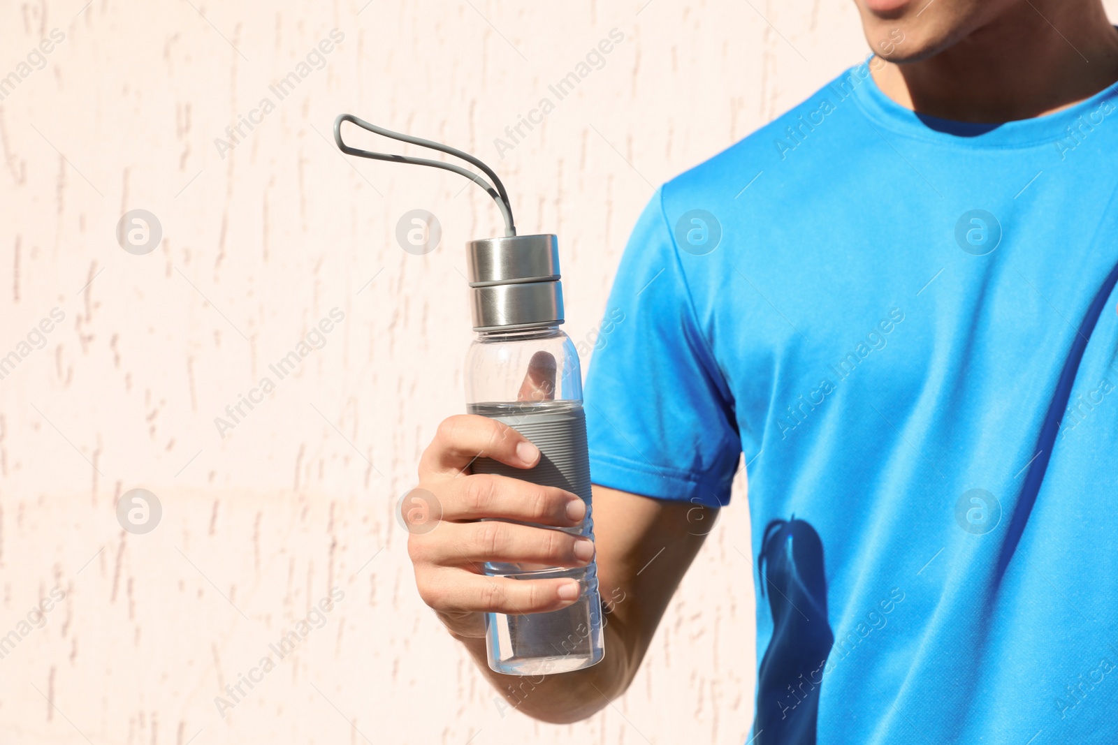 Photo of Sporty man holding bottle of water near wall outdoors on sunny day, closeup. Space for text