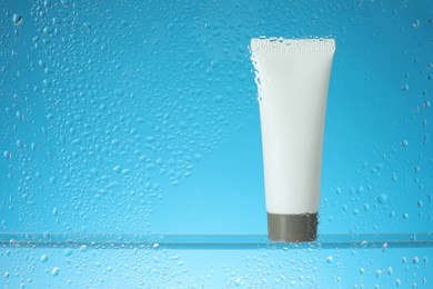 Tube with moisturizing cream on light blue background, view through wet glass. Space for text