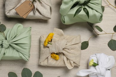 Photo of Furoshiki technique. Many gifts packed in fabric, flowers and eucalyptus leaves on wooden table, flat lay