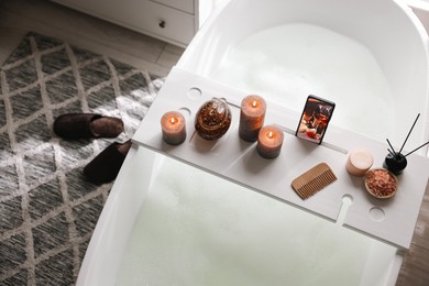 Photo of White wooden tray with smartphone, burning candles and spa products on bathtub in bathroom, above view