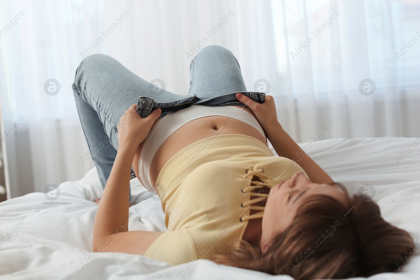 Photo of Young woman struggling to squeeze into tight jeans while lying on bed at home