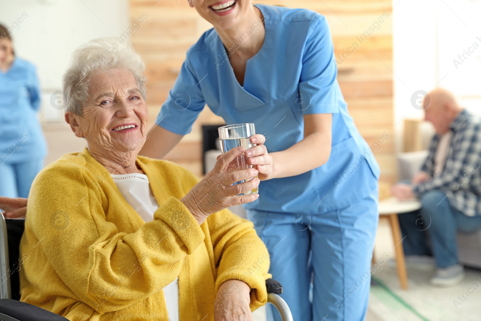 Photo of Nurse giving glass of water to elderly woman in wheelchair at retirement home. Assisting senior people