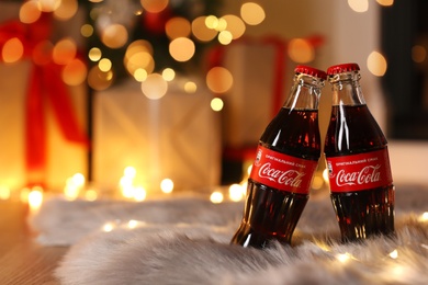 Photo of MYKOLAIV, UKRAINE - JANUARY 18, 2021: Coca-Cola bottles and fairy lights on fur rug in room, space for text