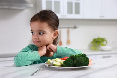 Photo of Cute little girl refusing to eat vegetables in kitchen
