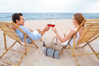 Happy young couple with glasses of wine sitting on deck chairs at sea beach