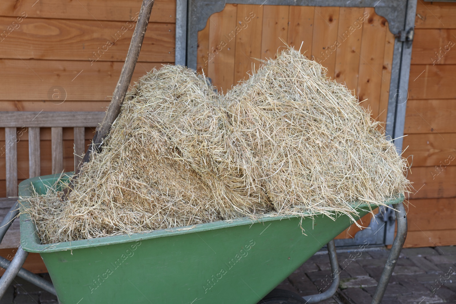 Photo of Wheelbarrow with hay near wooden stable outdoors, closeup