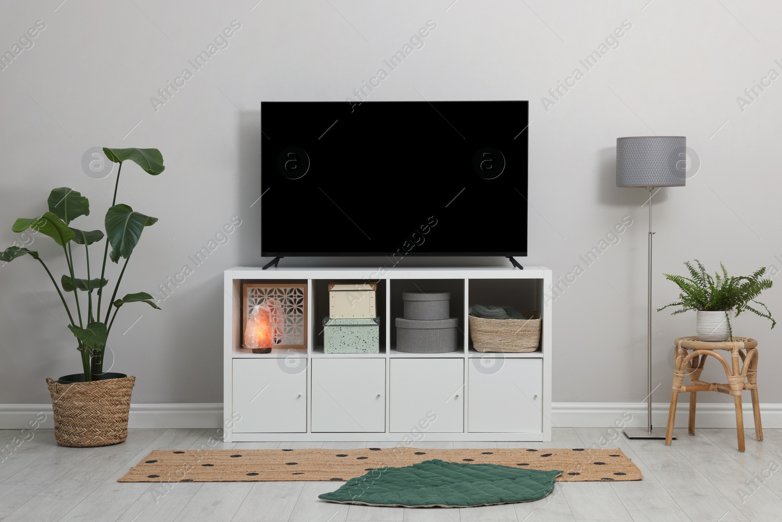 Photo of Modern TV on cabinet, lamp and beautiful houseplants near white wall indoors. Interior design