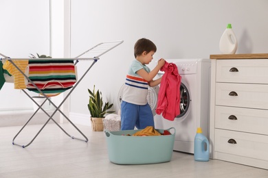 Cute little boy with laundry near washing machine and clothes drying rack indoors