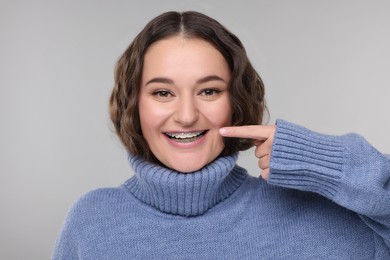 Happy woman in warm sweater pointing at braces on her teeth against grey background