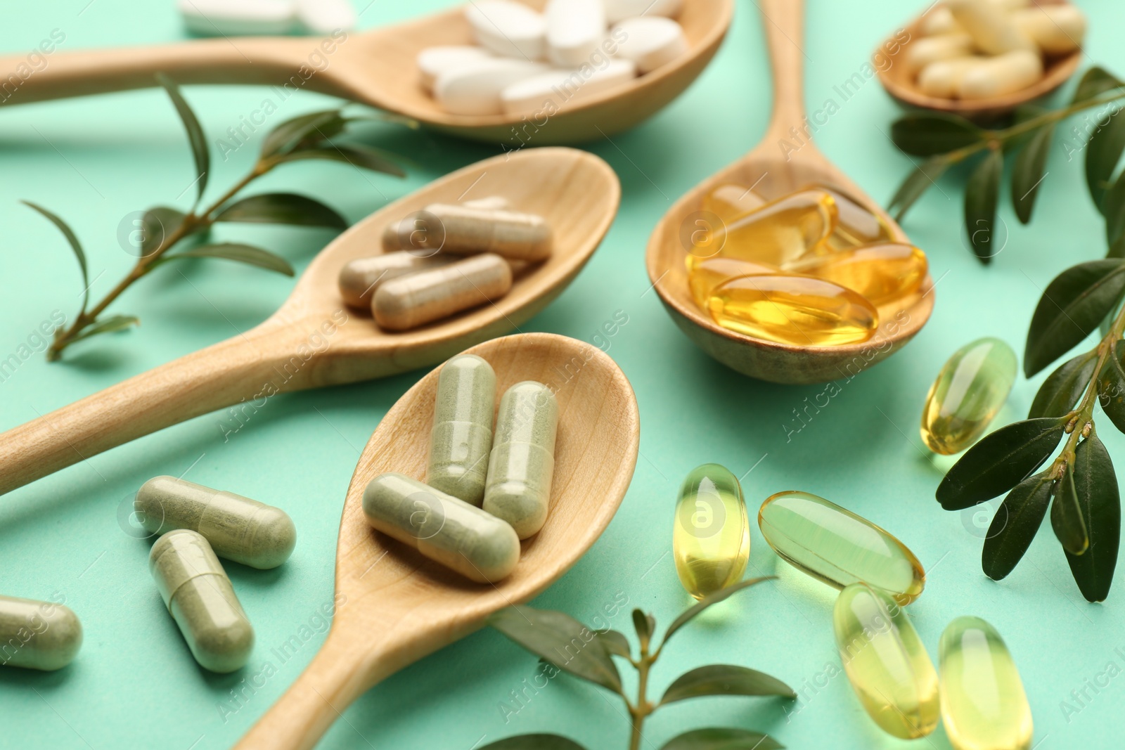 Photo of Different vitamin capsules in spoons and leaves on turquoise background, closeup