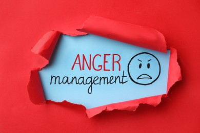 Photo of Text Anger Management and drawn angry face on light blue background, view through torn red paper