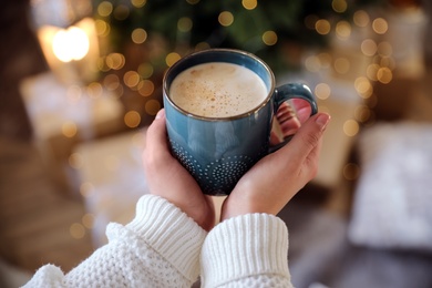 Photo of Woman holding cup of cocoa near Christmas tree indoors, closeup