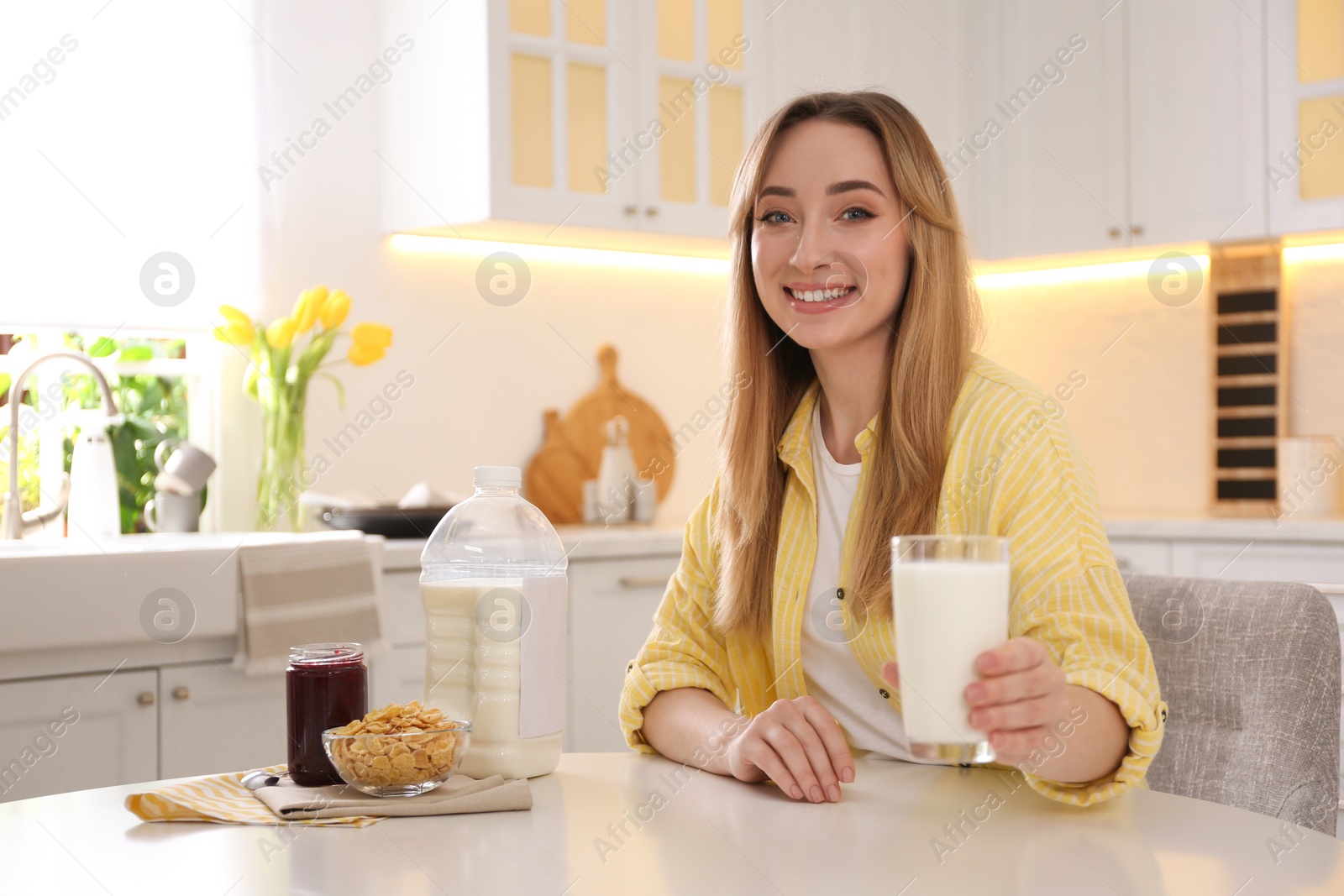 Photo of Young woman with gallon bottle of milk, glass and breakfast cereal at white table in kitchen