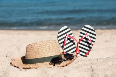 Photo of Striped flip flops, straw hat and sunglasses on sandy beach near sea