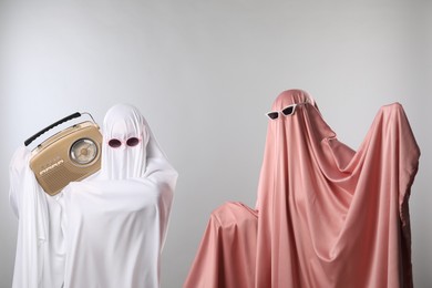 Photo of Glamorous ghosts. Women in color sheets and sunglasses with retro radio receiver on light grey background
