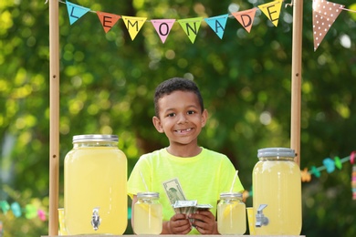 Cute little African-American boy with money at lemonade stand in park. Summer refreshing natural drink