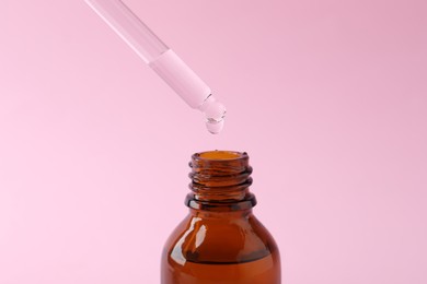 Dropping cosmetic oil from pipette into bottle on pink background, closeup
