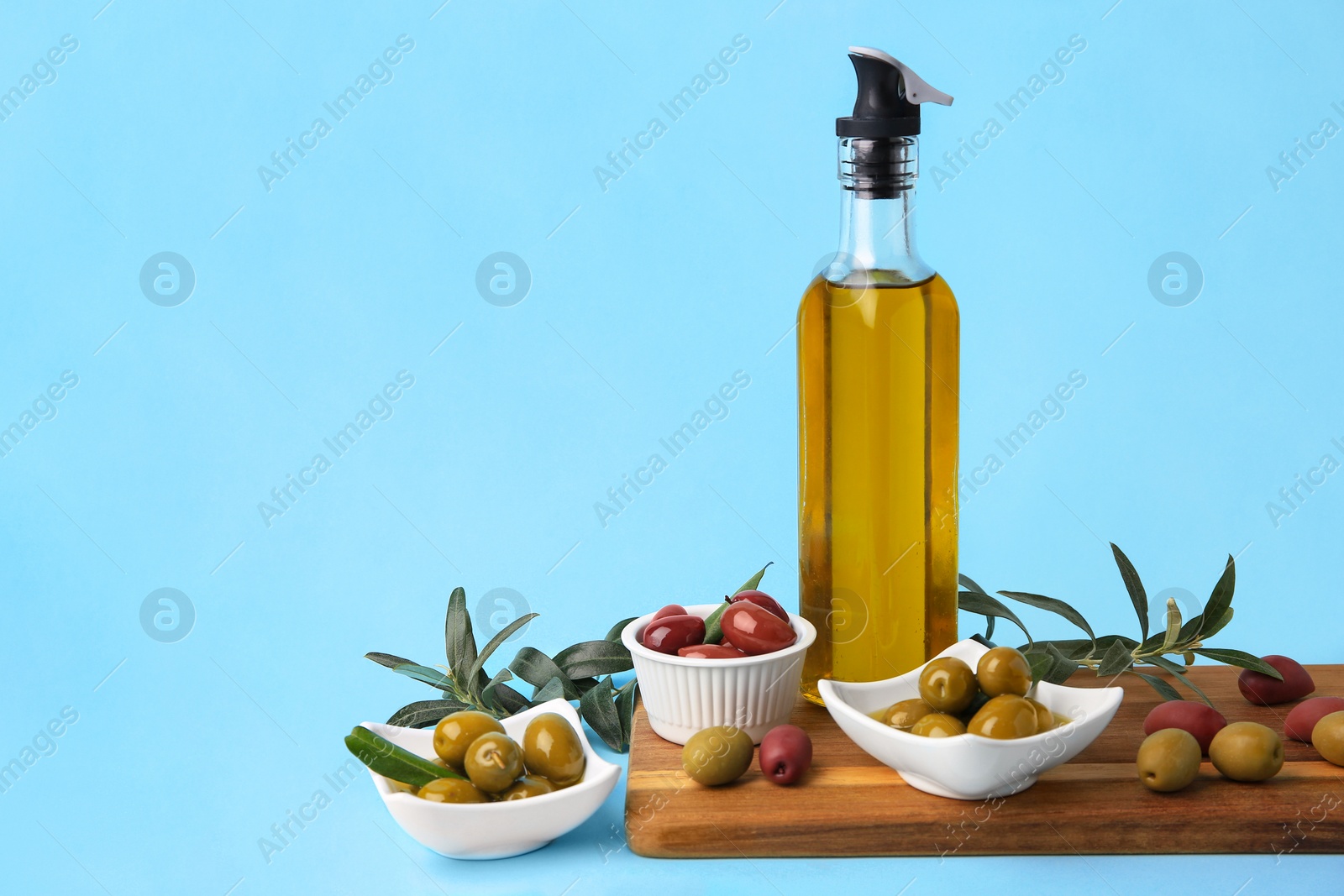 Photo of Bottle of oil, olives and tree twigs on light blue background, space for text