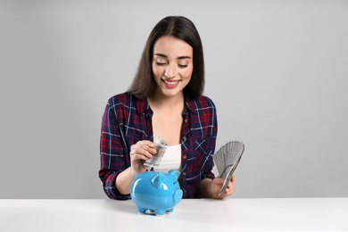 Young woman putting money into piggy bank at table on light grey background