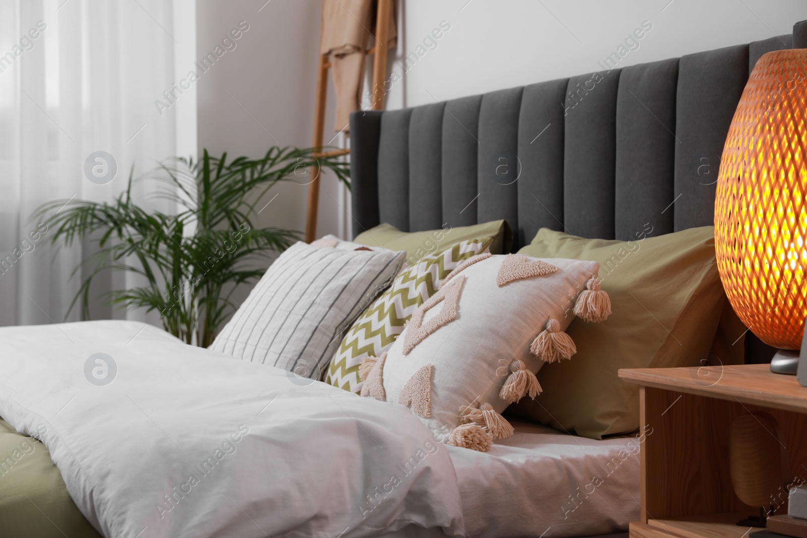 Photo of Comfortable bed with cushions, lamp on wooden bedside table in room. Stylish interior