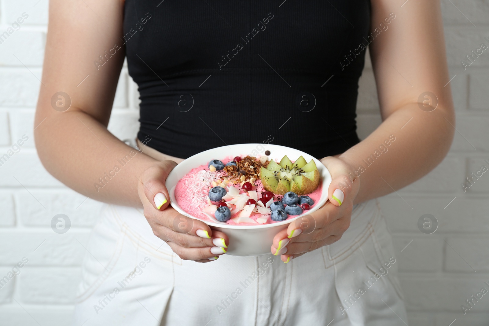 Photo of Woman holding tasty smoothie bowl with fresh kiwi fruit, berries and granola near white brick wall, closeup