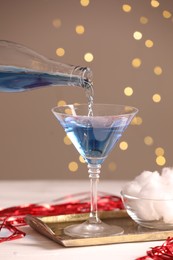 Photo of Cocktail making. Pouring tasty alcoholic drink in glass at white wooden table against beige background with blurred lights, closeup