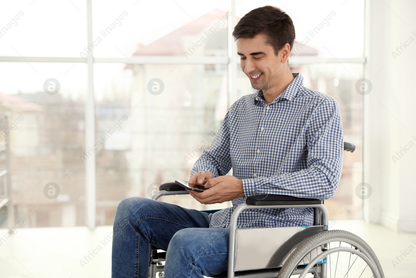 Photo of Young man in wheelchair using mobile phone near window indoors