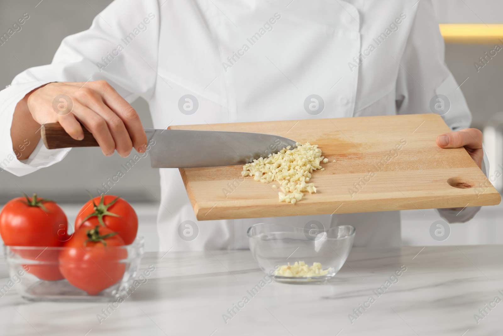 Photo of Professional chef putting cut garlic into bowl at white marble table indoors, closeup