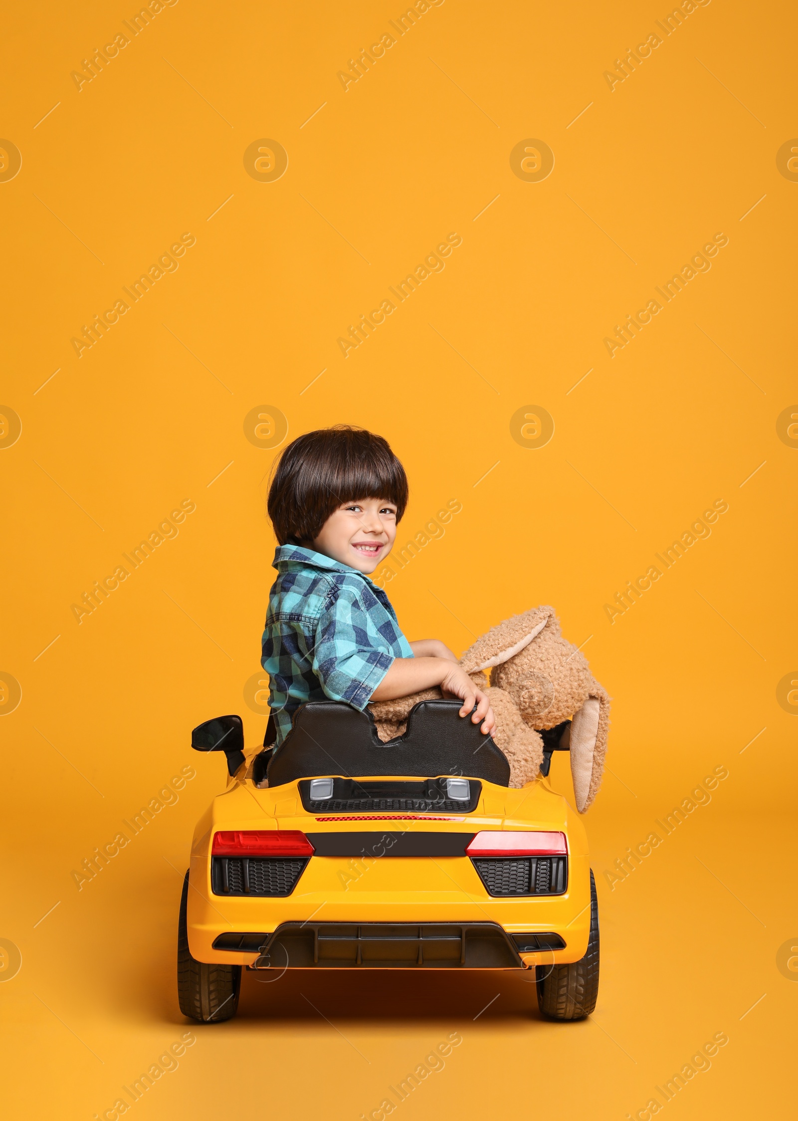 Photo of Cute little boy with toy bunny driving children's car on yellow background