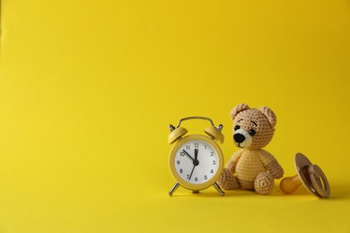 Photo of Alarm clock, toy bear and baby dummy on yellow background, space for text. Time to give birth