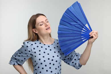 Beautiful woman waving blue hand fan to cool herself on light grey background