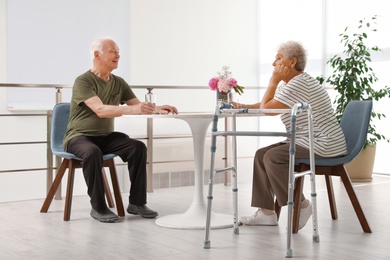 Photo of Man and his wife with walking frame sitting at table indoors