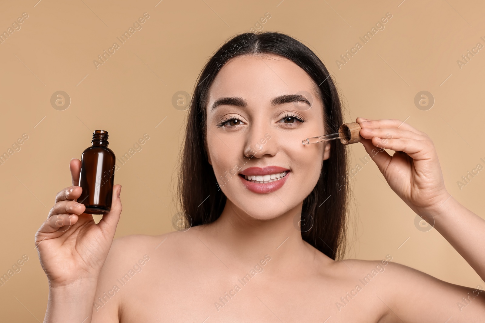 Photo of Happy young woman with bottle applying essential oil onto face on beige background