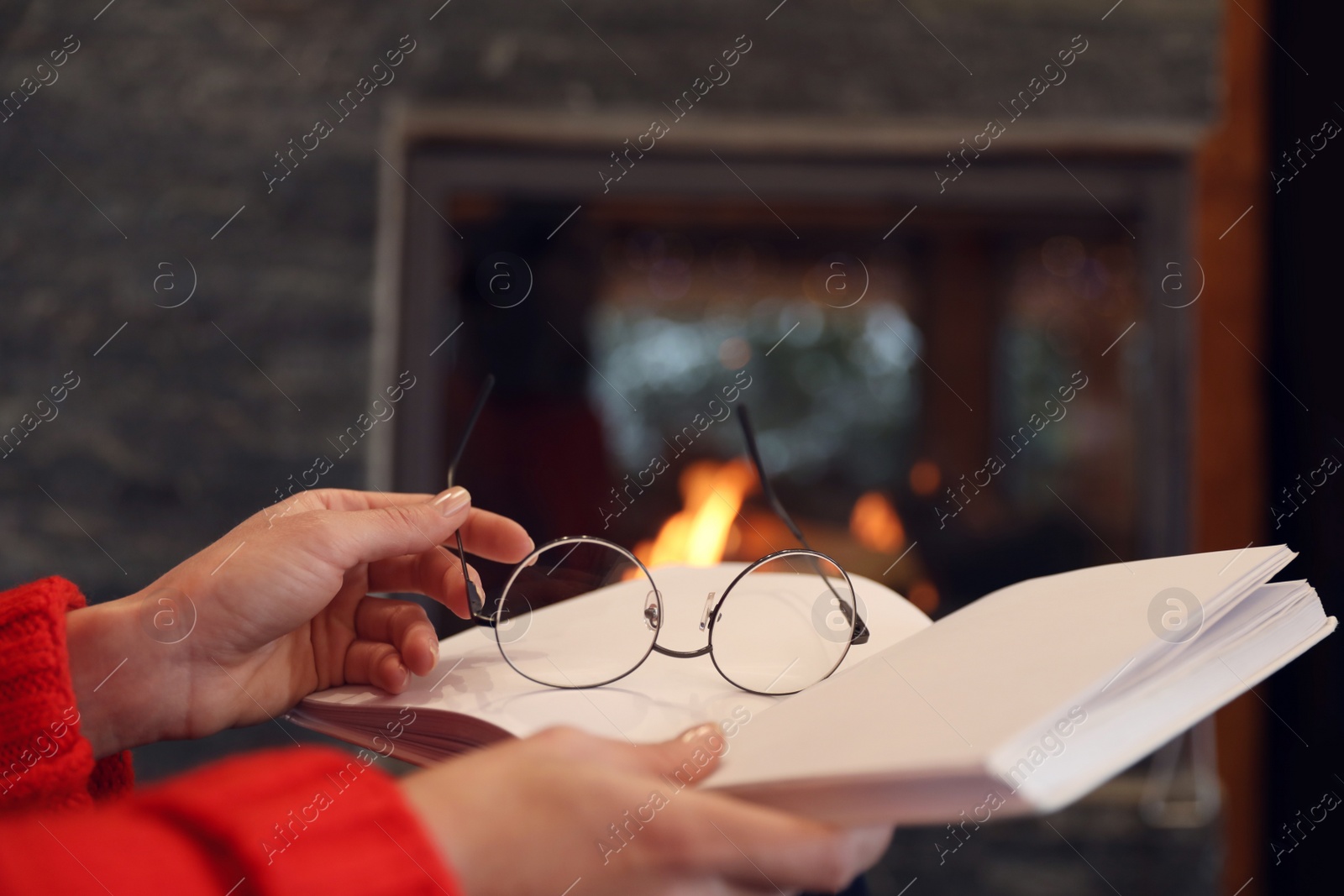 Photo of Woman with glasses reading book near burning fireplace, closeup