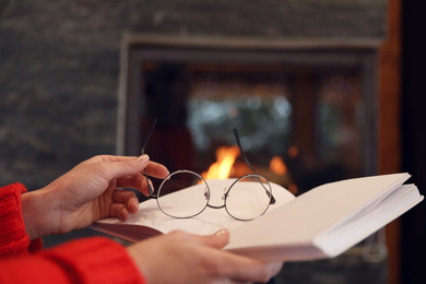 Woman with glasses reading book near burning fireplace, closeup