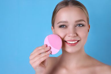Young woman washing face with cleansing brush on light blue background. Cosmetic product
