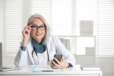 Photo of Mature female doctor with smartphone at table in office