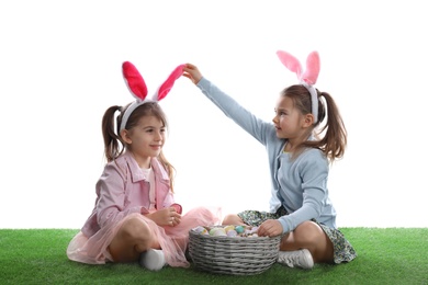 Adorable little girls with bunny ears and wicker basket full of Easter eggs on green grass against white background