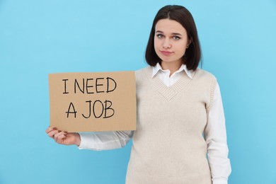 Young unemployed woman holding sign with phrase I Need A job on light blue background