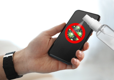 Image of Sanitizing mobile devices during coronavirus outbreak. Antiseptic spray and man with smartphone on light background, closeup