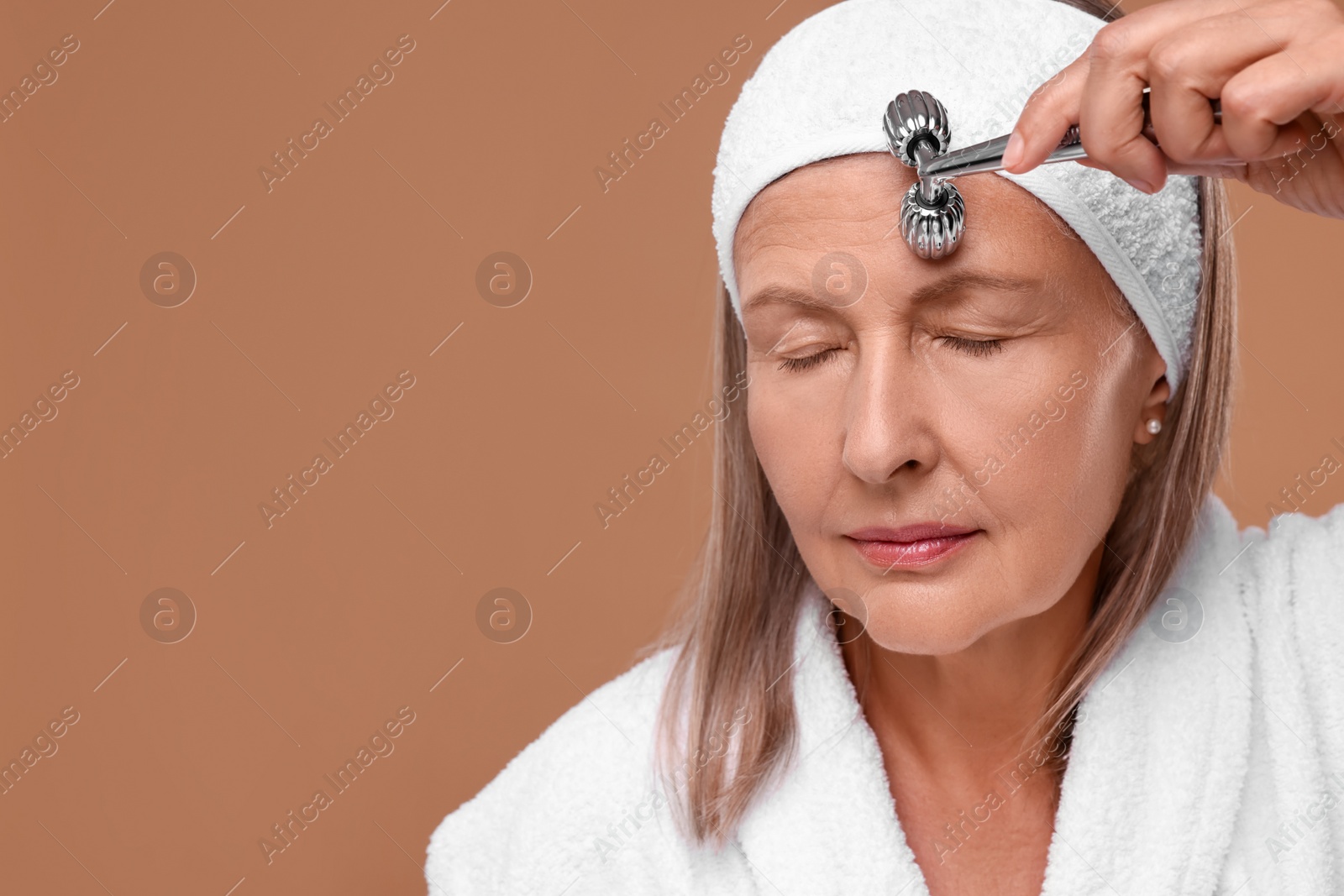 Photo of Woman massaging her face with metal roller on brown background. Space for text