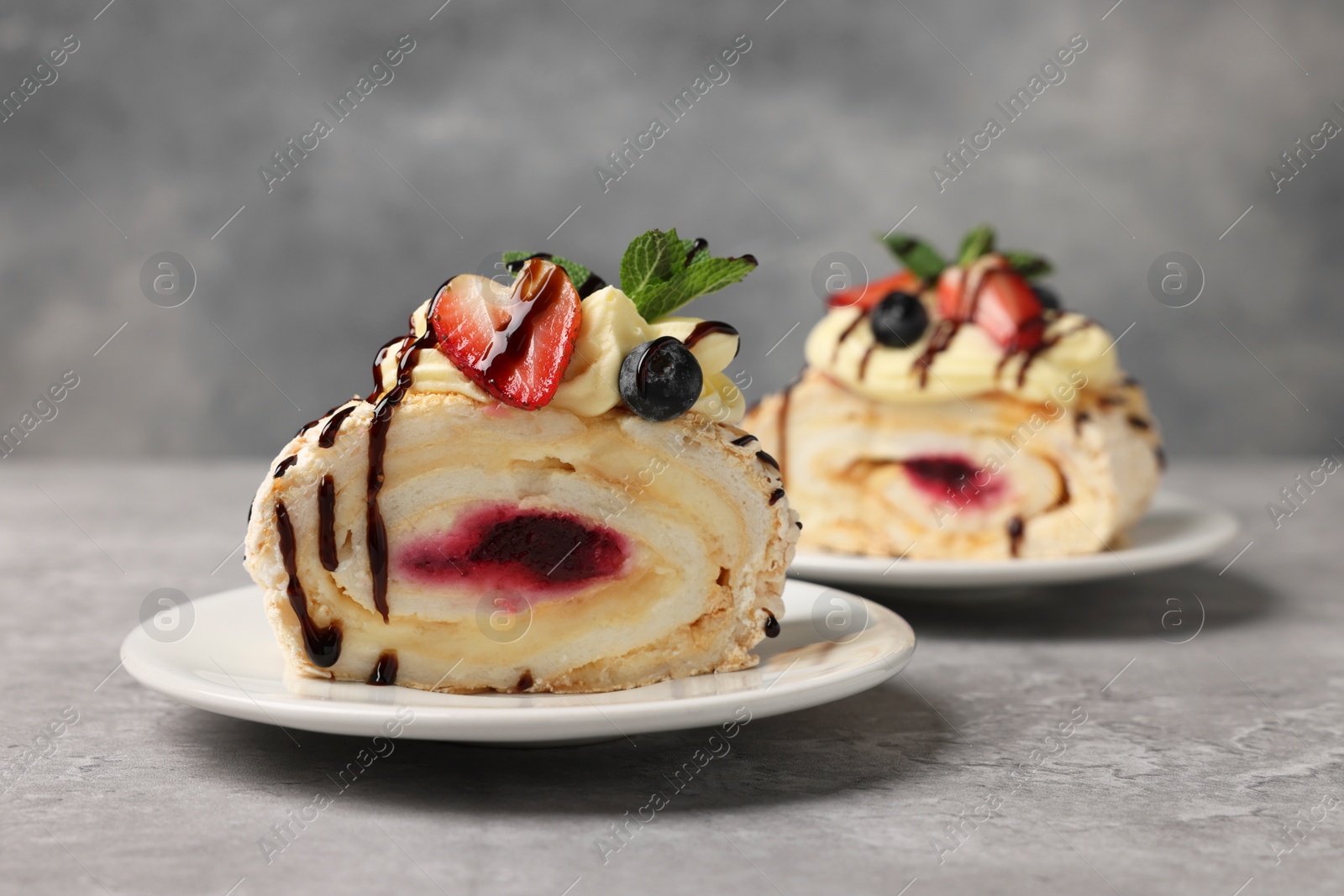 Photo of Pieces of tasty meringue roll with jam, cream, strawberry, blueberry and mint on light grey table, closeup