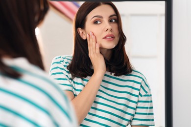 Photo of Young woman looking in mirror and touching her face indoors. Hormonal disorders